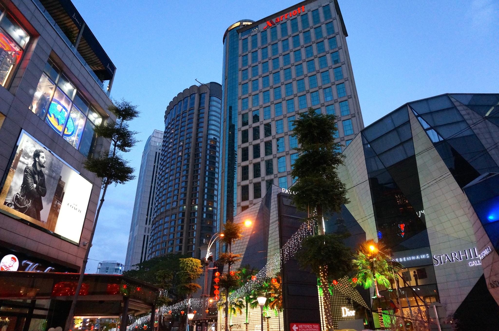Отель Jw Marriott Куала-Лумпур Экстерьер фото The photo shows a modern urban scene during twilight. There are several tall buildings, including a notable high-rise with a glass facade, suggesting a commercial area. The architecture appears contemporary, with a mix of reflective surfaces and illu