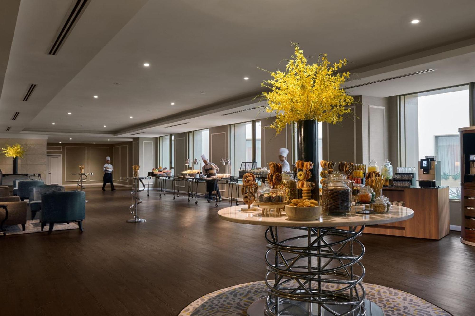 Отель Jw Marriott Куала-Лумпур Экстерьер фото The photo shows a modern, elegant dining area, likely set up for a buffet or catering event. In the center, there is a large, round table with a decorative arrangement of yellow flowers, accompanied by various jars and displays of food items. Several