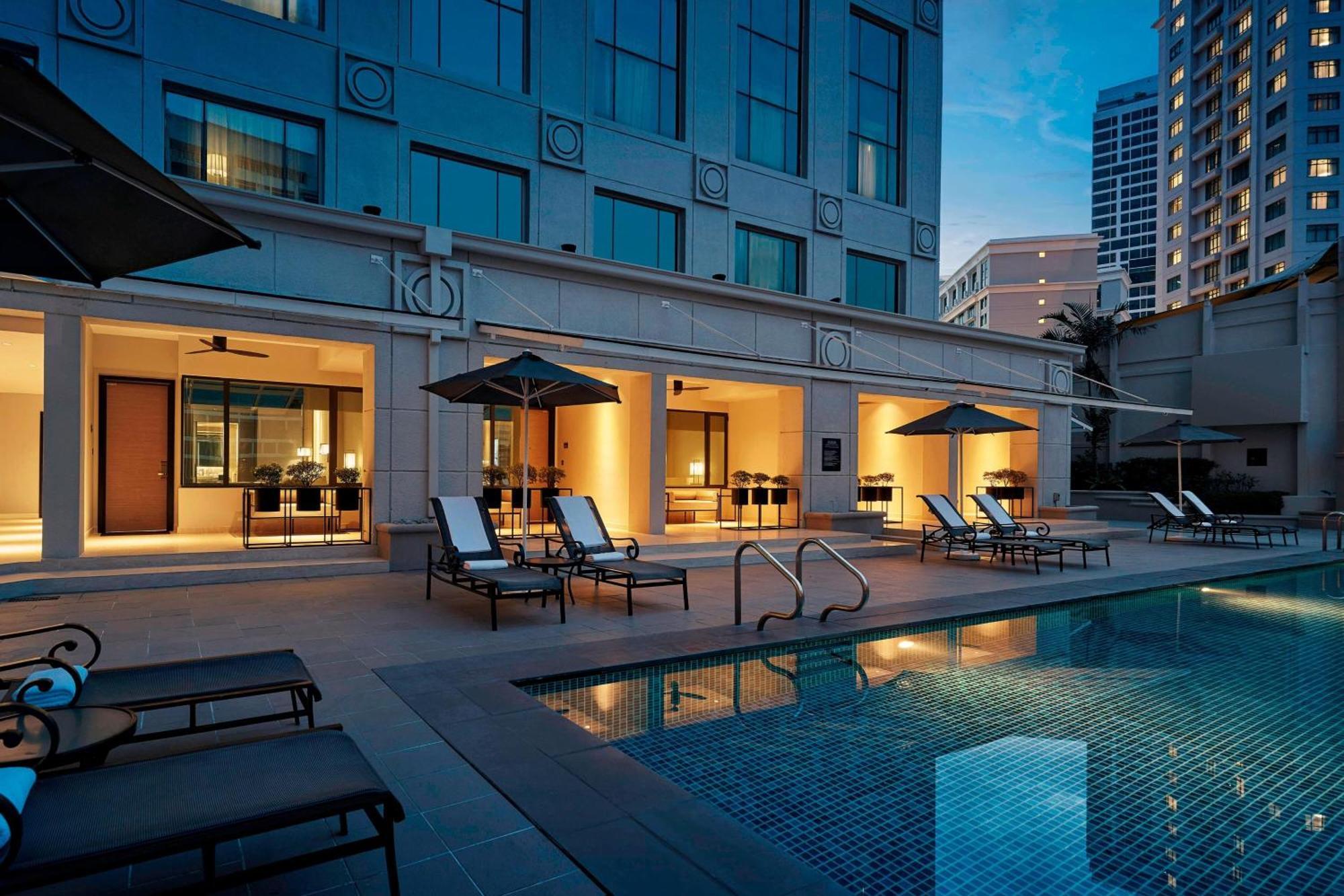 Отель Jw Marriott Куала-Лумпур Экстерьер фото The photo depicts a luxurious hotel pool area. The swimming pool features clear water and is surrounded by several lounge chairs under sun umbrellas. In the background, there are large glass windows of the hotel, showing illuminated spaces inside, li