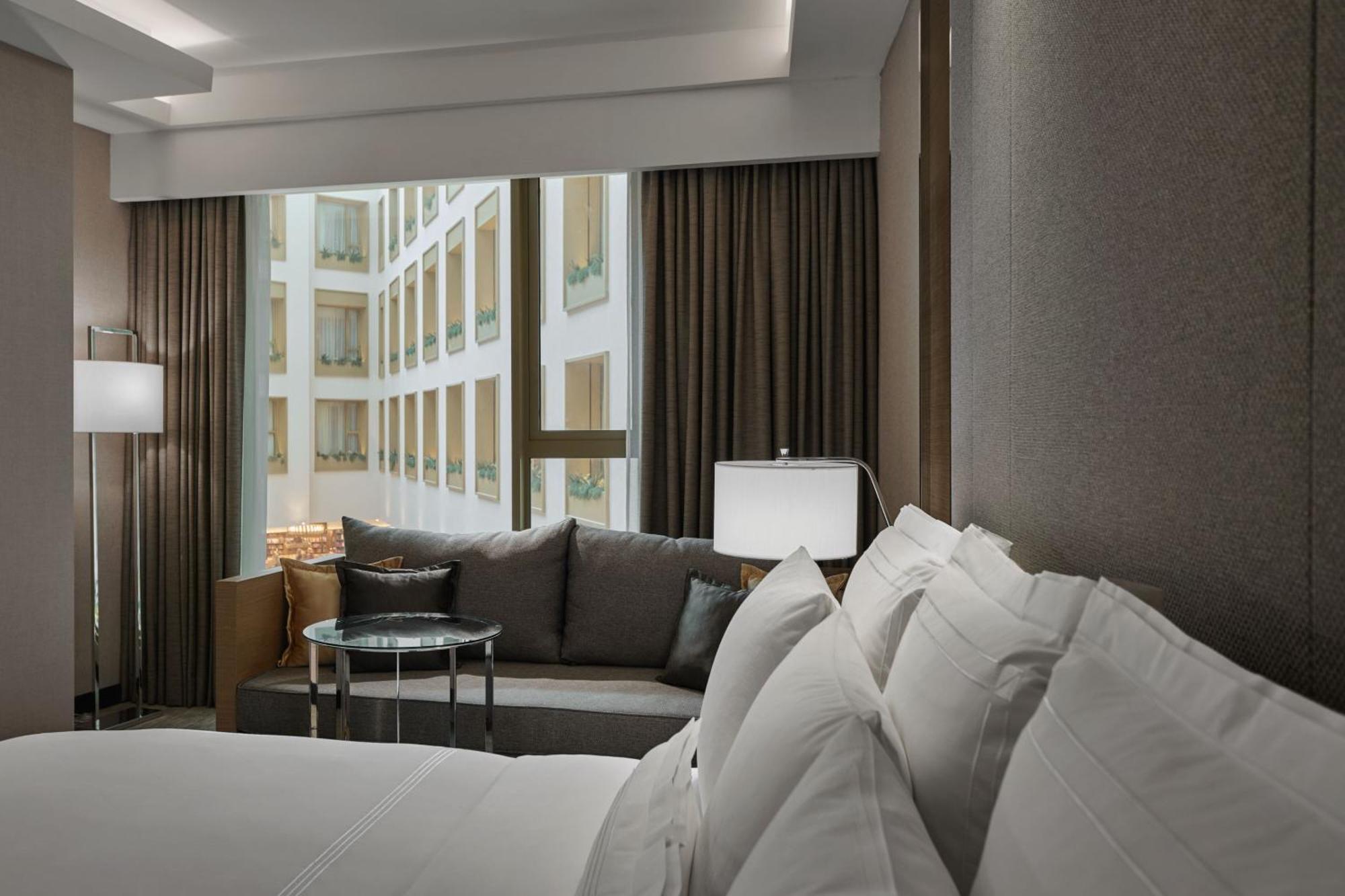 Отель Jw Marriott Куала-Лумпур Экстерьер фото The photo features a modern hotel room. In the foreground, there is a large bed with crisp, white linens and multiple pillows. To the side, there is a cozy seating area with a gray sofa and a round glass table. A lamp is situated next to the sofa, pr