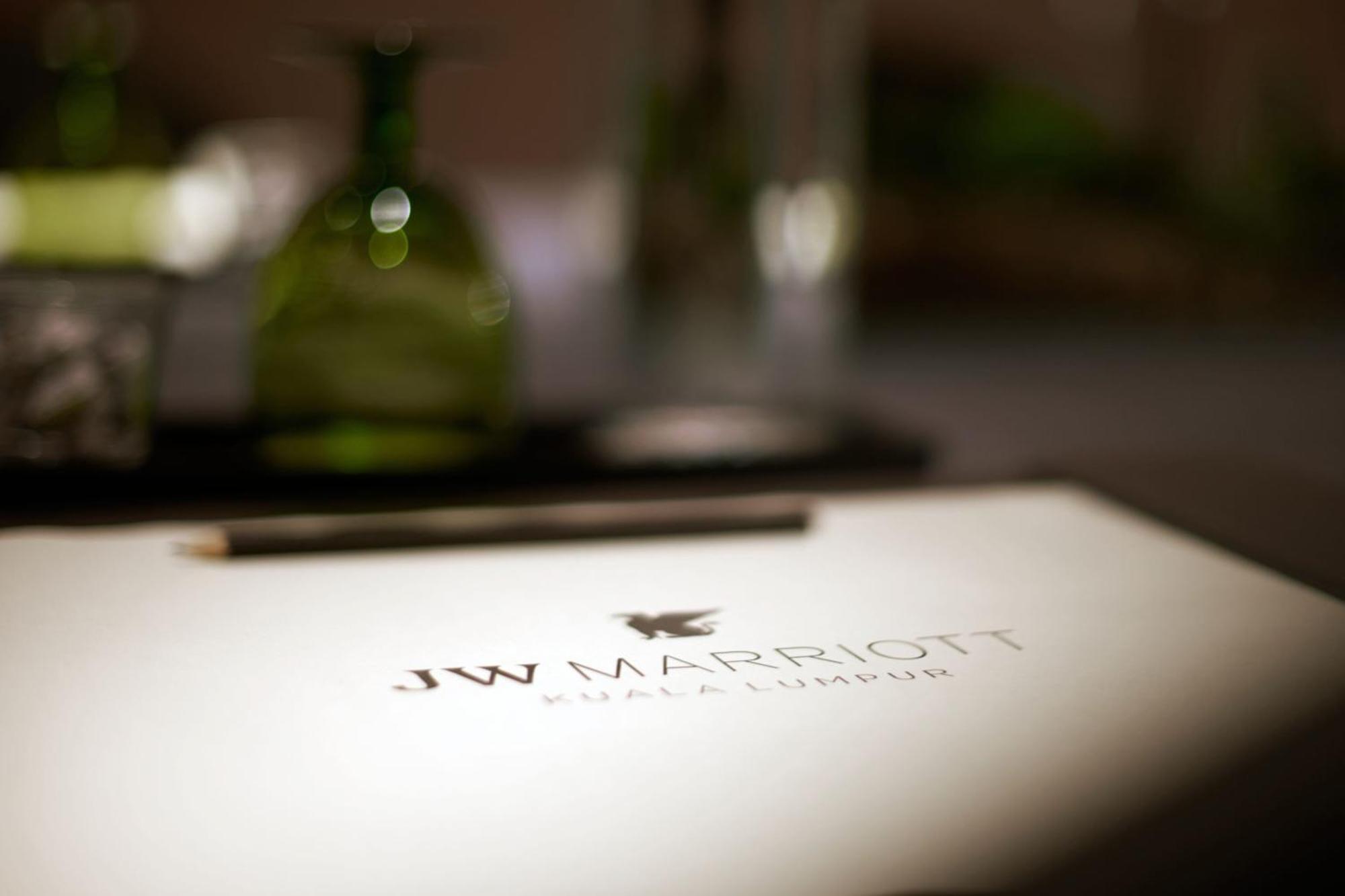 Отель Jw Marriott Куала-Лумпур Экстерьер фото The photo shows a close-up of a table setting with a focus on a menu or notepad featuring the logo of "JW Marriott Kuala Lumpur." The logo is prominently displayed in a refined, elegant font. In the background, there are green decorative bottles and 