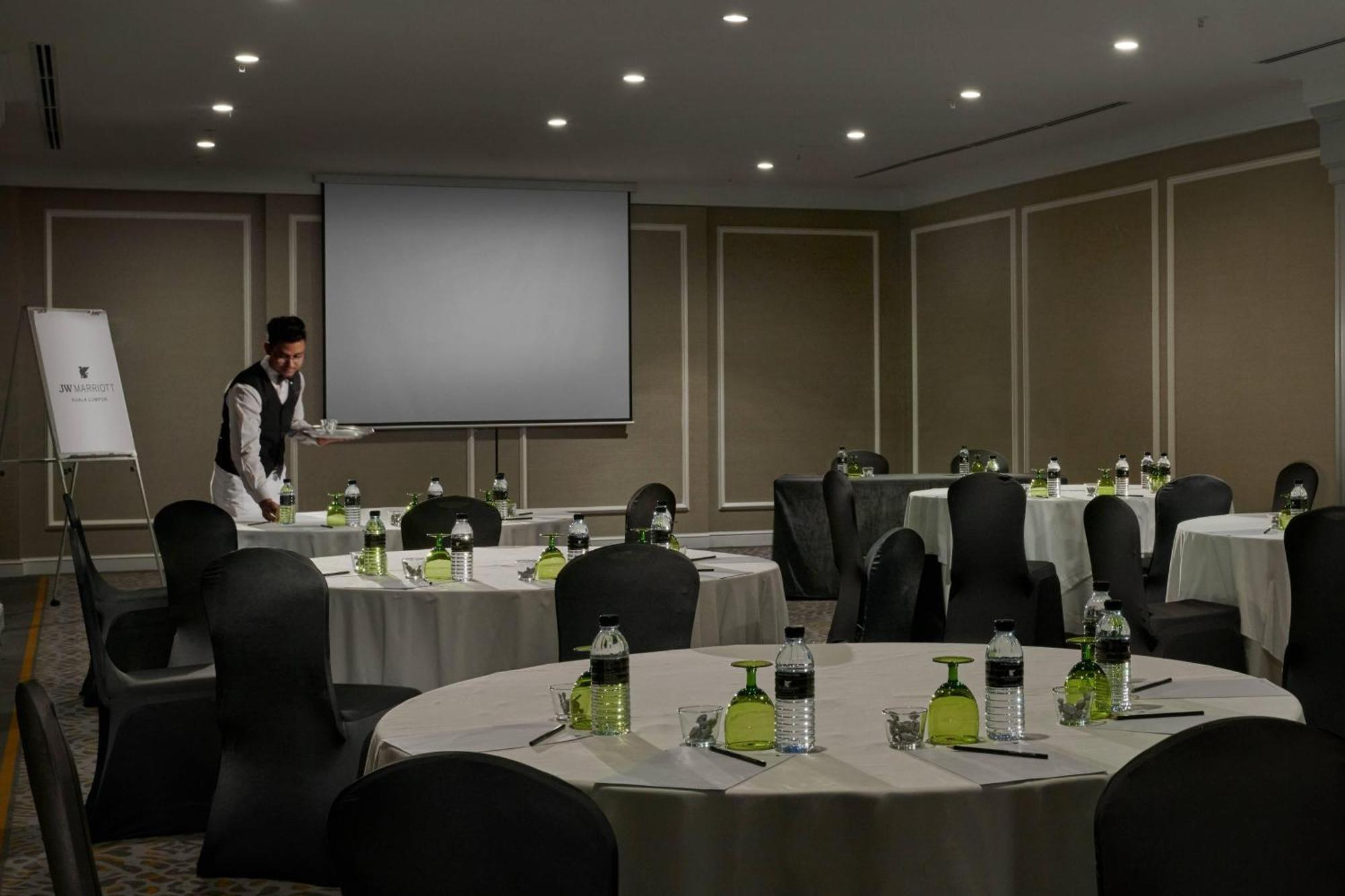 Отель Jw Marriott Куала-Лумпур Экстерьер фото The photo shows a conference or meeting room setup. There are several round tables covered with light-colored tablecloths, each surrounded by black chairs. On the tables, there are green table settings, including small bottles and drinkware. A projec