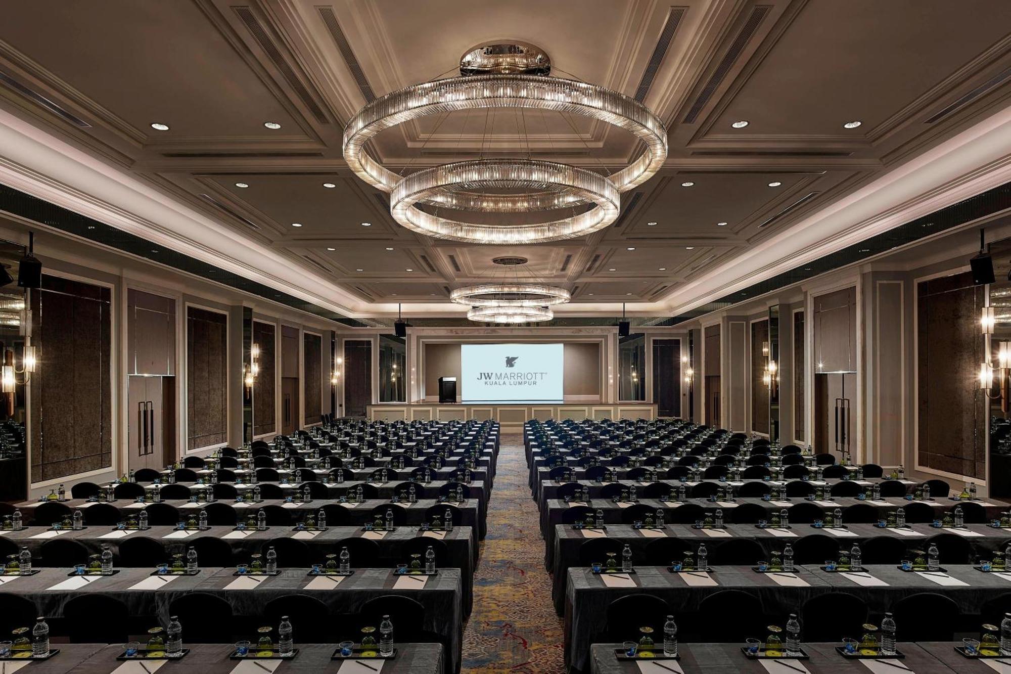 Отель Jw Marriott Куала-Лумпур Экстерьер фото The photo shows a large conference room set up for a meeting or event. There are numerous rows of tables arranged in a classroom or theater style, with chairs facing the front where a large screen is set up, likely for presentations. The room has ele
