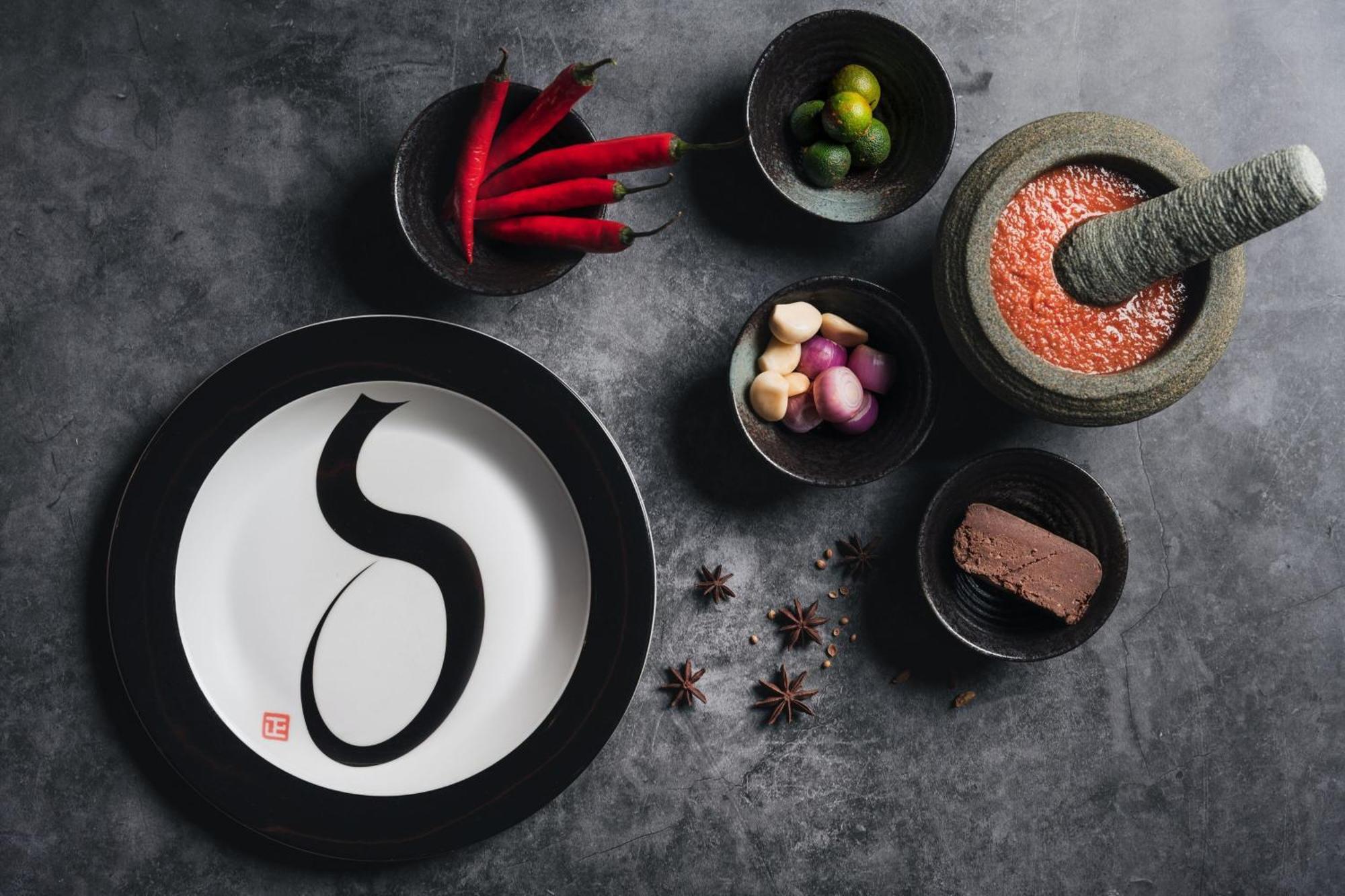 Отель Jw Marriott Куала-Лумпур Экстерьер фото The photo depicts an artistic arrangement of food items on a textured gray surface. In the center, there is a white plate with a black rim featuring a stylized black letter or character. Surrounding the plate are several small bowls filled with vario