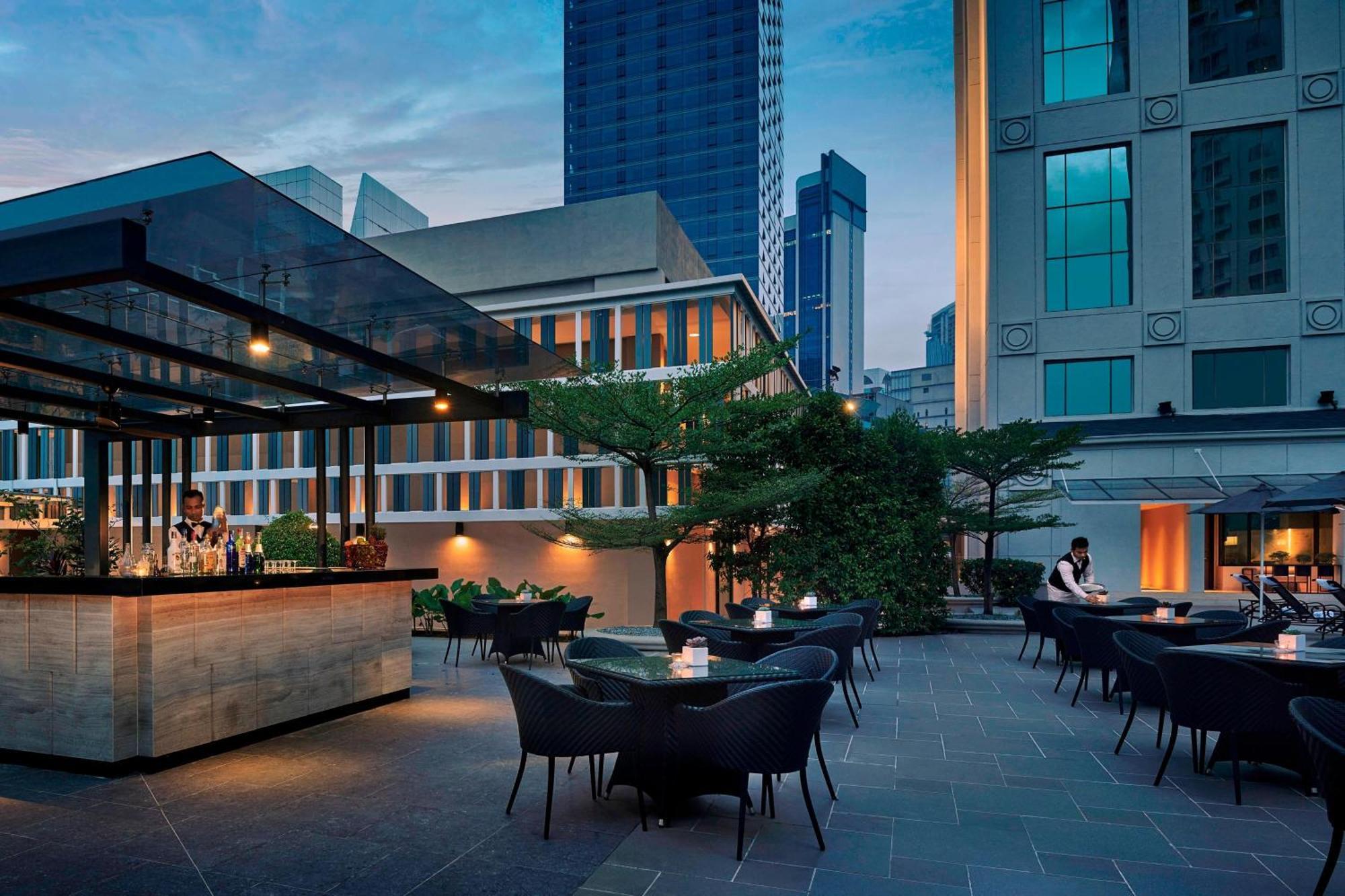 Отель Jw Marriott Куала-Лумпур Экстерьер фото The photo depicts a modern outdoor terrace or rooftop bar. It features a stylish bar area with an elegant glass structure, set against a backdrop of tall buildings. There are several tables with dark, woven chairs, arranged for guests to sit. The atm
